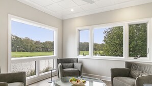 A modern living room with a glass coffee table, grey sofas, and large windows offering a view of a green landscape. - Views and location, great villa in Turtle Point (Kiawah Island)