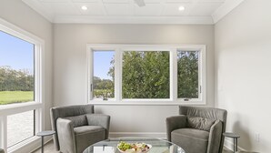 A modern living room with a glass coffee table, two grey armchairs, and a view of a green landscape through large windows. - Views and location, great villa in Turtle Point (Kiawah Island)