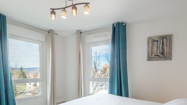 Un dormitorio con una cama grande, dos ventanas con cortinas, una araña y un cuadro en la pared.