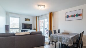 Un salón moderno con un sofá gris, una mesa de comedor con cuatro sillas, un televisor de pantalla plana y una pintura de un paisaje urbano en la pared.