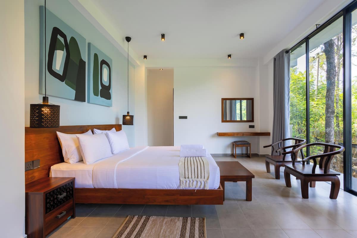 A modern bedroom with a large bed, bedside tables, a small seating area, and a view of trees through large windows.