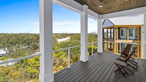 A covered outdoor deck with wooden flooring and white railings, overlooking a lush landscape and a distant building. - The Boat House at Kiva Dunes (Gulf Shores)