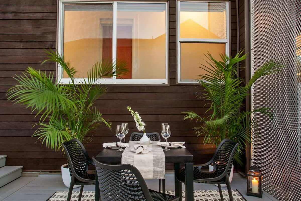 An outdoor dining area with a table set for two, surrounded by chairs, and two potted plants.