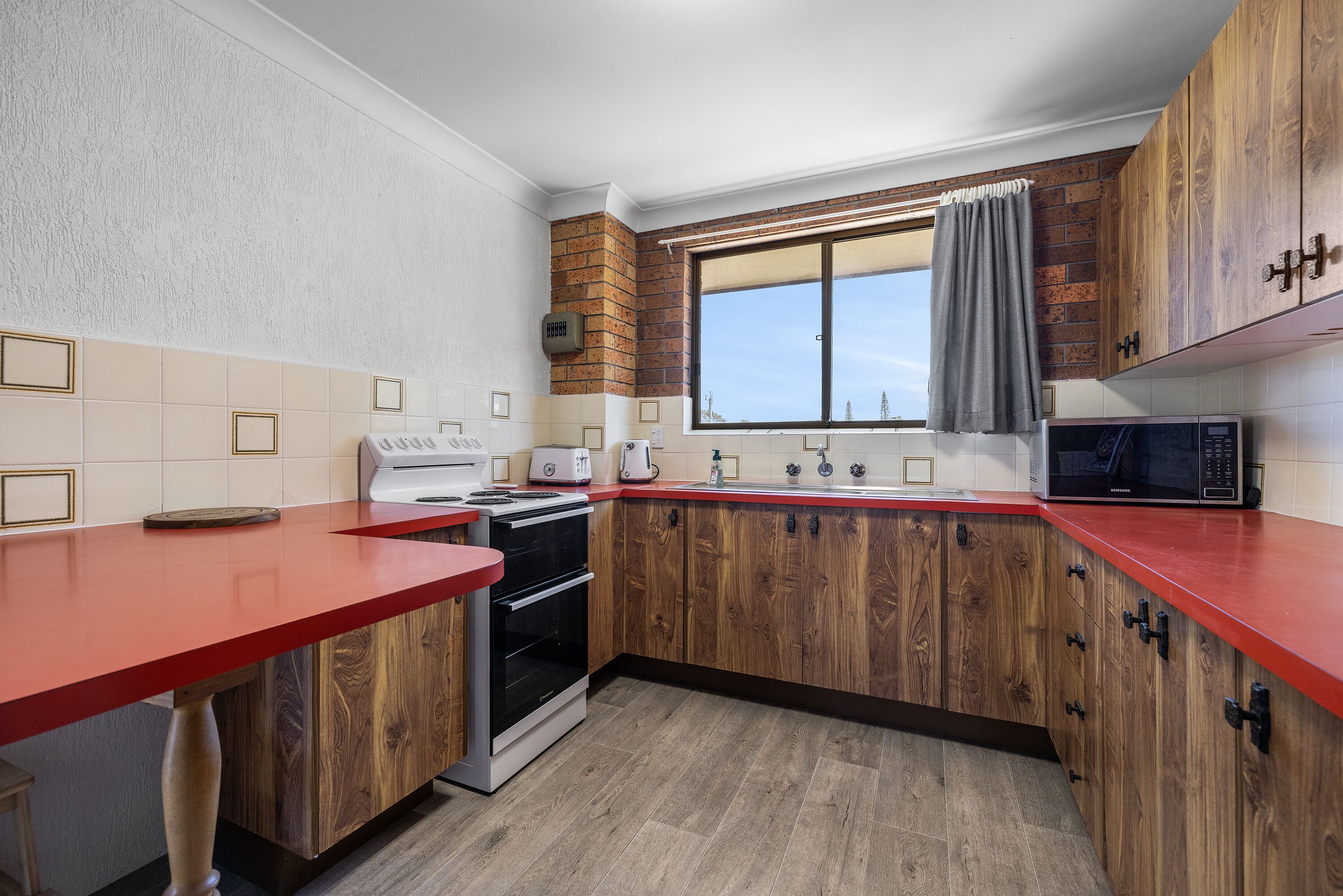 A kitchen with wooden cabinets, a red countertop, a built-in oven, and a window with a curtain.