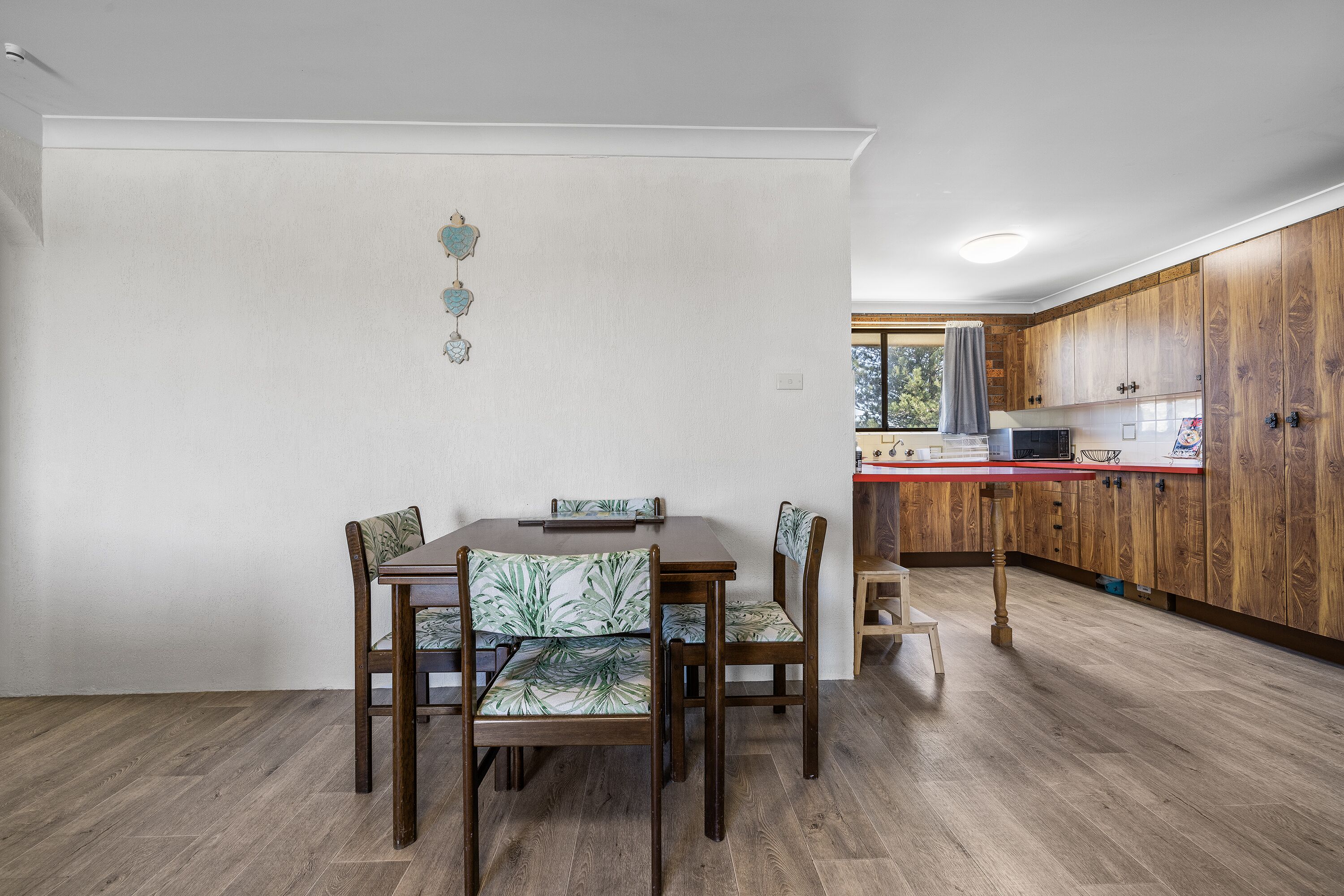 A dining area with a wooden table and chairs, a kitchen with wooden cabinets, and a window with a view of trees.