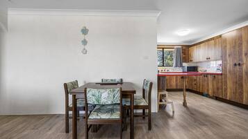 A dining area with a wooden table and chairs, a kitchen with wooden cabinets, and a window with a view of trees.