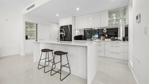 A modern kitchen with white cabinetry, a central island with stools, and a glass-fronted pantry. - Paradise on Gregory G04/21 Gregory St SWR (South West Rocks)