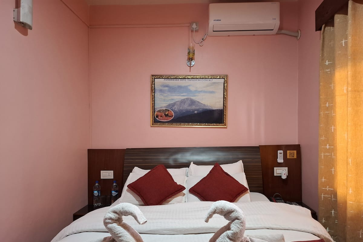A hotel room with a bed, pillows, a wall-mounted air conditioner, a framed picture, and a bedside table with water bottles.