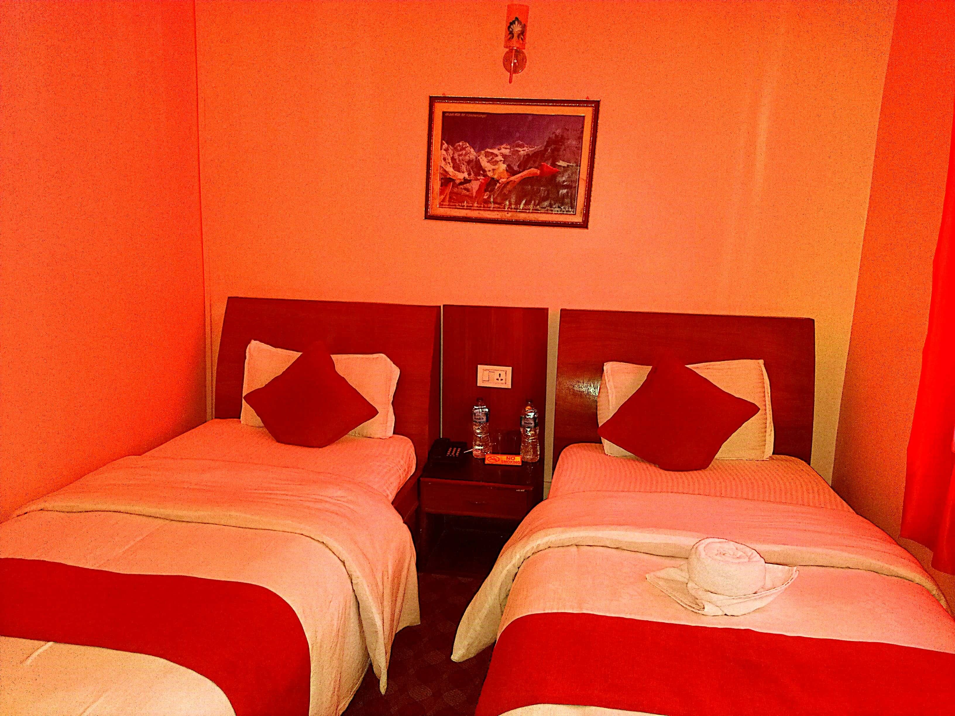 A hotel room with two beds, each with red bedding and pillows, a nightstand with a phone and a bottle, and a framed picture on the wall.
