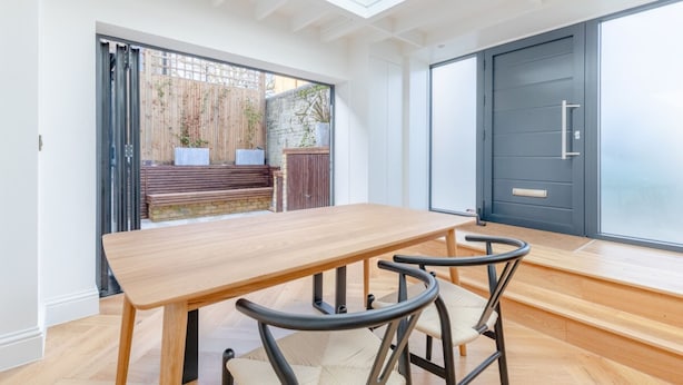 A modern dining area with a wooden table and chairs, a sliding glass door leading to an outdoor space, and a dark-colored door. - Luxuriously Compact 1BD Eco Home Clapham Common! (London)