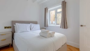 A neatly made bed with white linens and rolled towels, a bedside table with a lamp, and a window with curtains. - Luxuriously Compact 1BD Eco Home Clapham Common! (London)