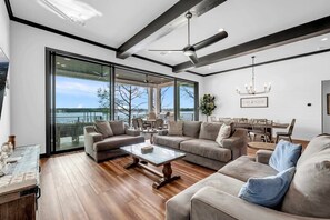 A spacious living room with a large window overlooking a body of water, a ceiling fan, and a chandelier.