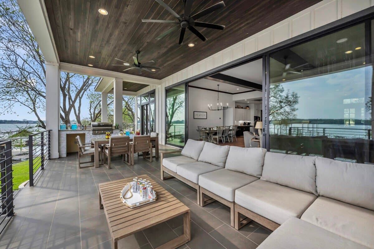 A modern outdoor living area with a wooden ceiling, ceiling fans, a large sofa, a dining table, and a kitchen area visible through glass doors.