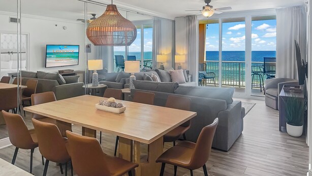 A spacious living area with a wooden dining table, comfortable seating, and a large TV screen. - Azure #613 Updated Top Floor 3bdrm Sleeps 10! Views Galore! (Fort Walton Beach)