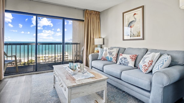 A living room with a view of the ocean, a sofa with decorative pillows, a coffee table, and a framed picture on the wall. - Vacations By The Sea at Ocean Dunes Tower 1 (Myrtle Beach)