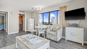 A modern living room with a dining area, a TV mounted on the wall, and a view of the city through large windows. - Vacations By The Sea at Ocean Dunes Tower 1 (Myrtle Beach)