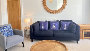 A living room with a dark blue sofa, a round wooden coffee table, a grey armchair with a blue patterned cushion, a wooden-framed mirror, and a small wooden side table with a lamp. - Sea Breeze-Cliff Terrace -Hunstanton. 3 Bedroom Apartment -Sea View  (Hunstanton)