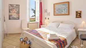 Un dormitorio con cama, ventana, silla, mesita de noche y lámpara.