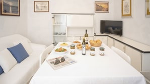 Un comedor con una mesa blanca dispuesta con comida, vino y copas. A la izquierda, un sofá blanco con un cojín azul.