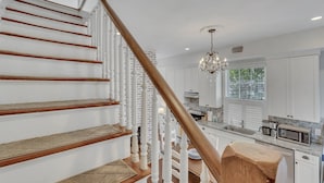 Un escalier avec des rampes blanches et des marches en bois menant à un espace cuisine doté de meubles blancs et d’un lustre.