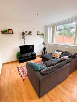 A living room with a dark grey sofa, a flat-screen TV, wooden flooring, and a window with a view of a brick wall. - Spacious 3 Bed Townhouse Garden Wifi Balham (London)
