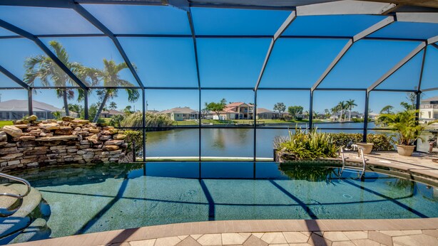 A pool with a clear view of a lake and houses in the background, surrounded by a metal fence and palm trees.