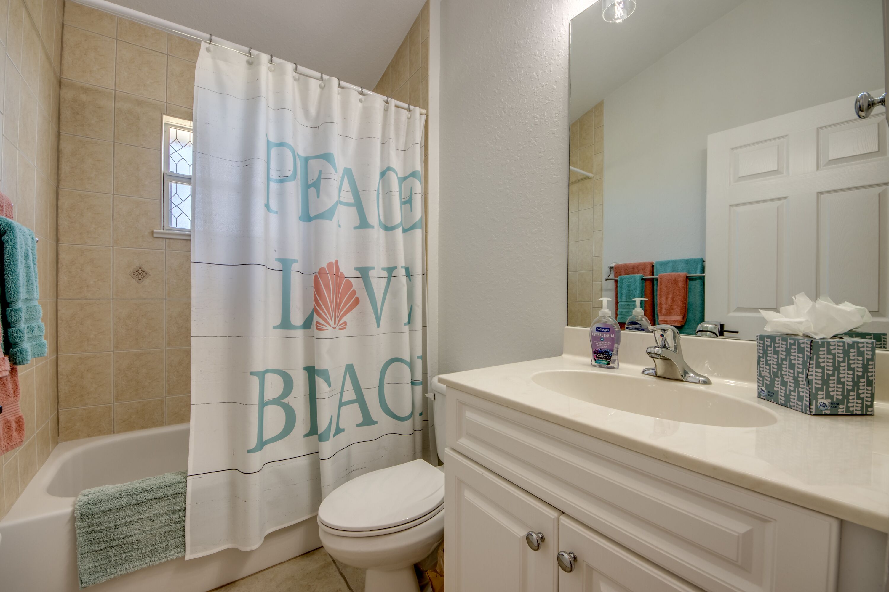 Un baño con cortina de ducha que dice "PEACE LOVE BEACH", un inodoro blanco, una bañera, un lavamanos con artículos de aseo y un espejo.