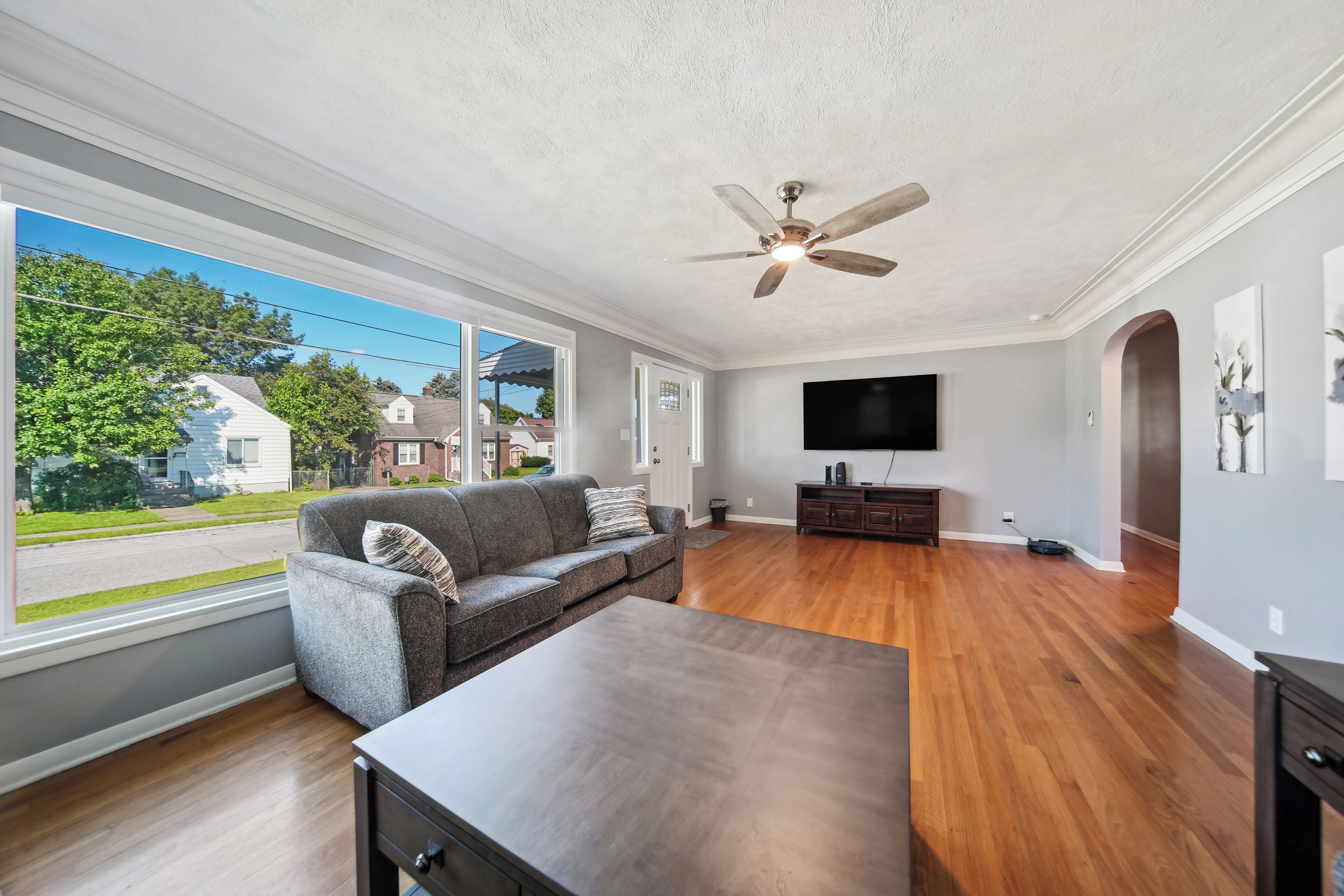 A spacious living room with hardwood floors, a gray sofa, a wooden coffee table, a ceiling fan, and a large window offering a view of the street and houses.