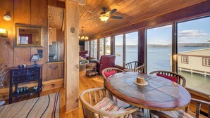 Ein Holzinterieur mit einem runden Tisch und Stühlen, großen Fenstern mit Blick auf das Wasser und einem Deckenventilator.