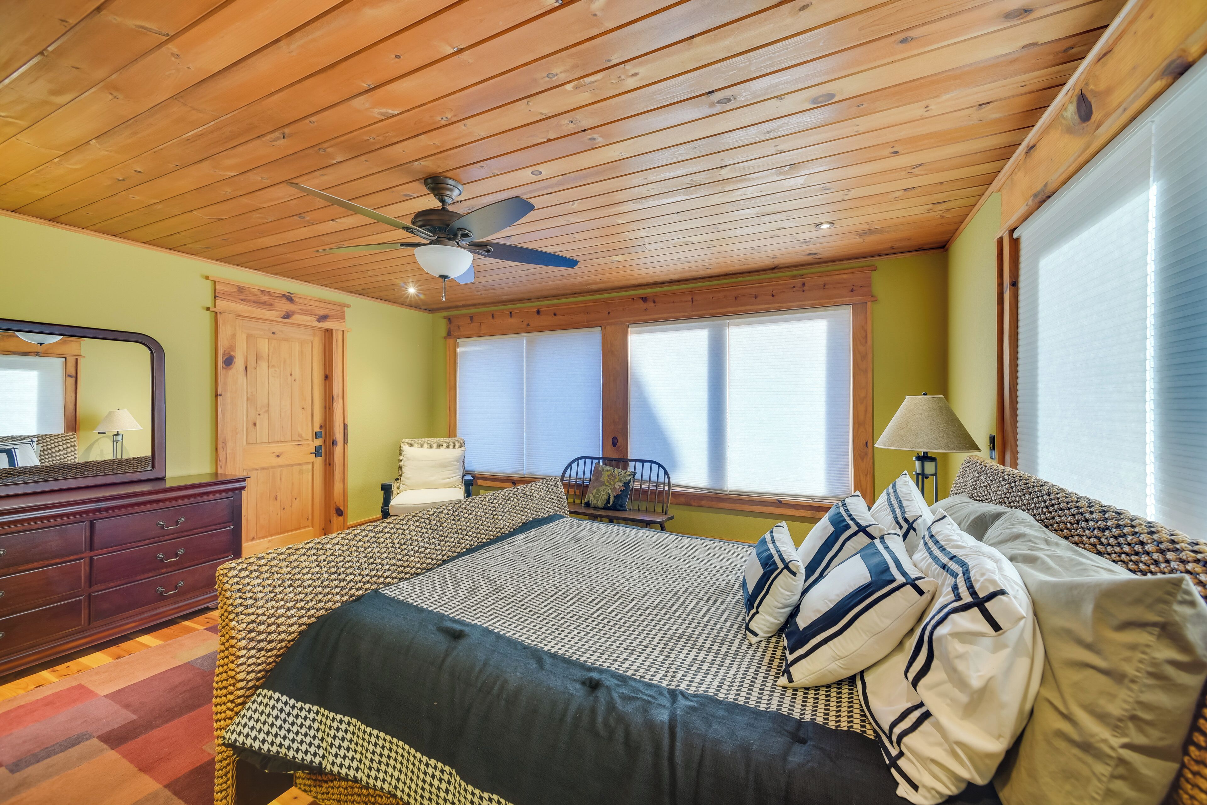 Une chambre avec un plafond en bois, un lit avec une literie à motifs, une commode et une fenêtre avec des stores.