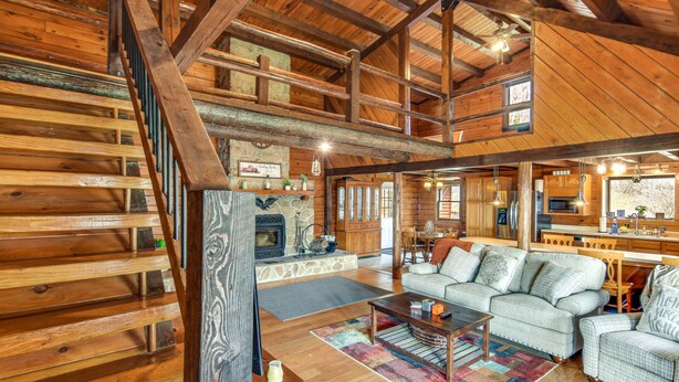 Un intérieur spacieux de chalet en bois, avec une cheminée, des poutres apparentes, un coin repas et un salon confortable doté d’un canapé et de fauteuils.