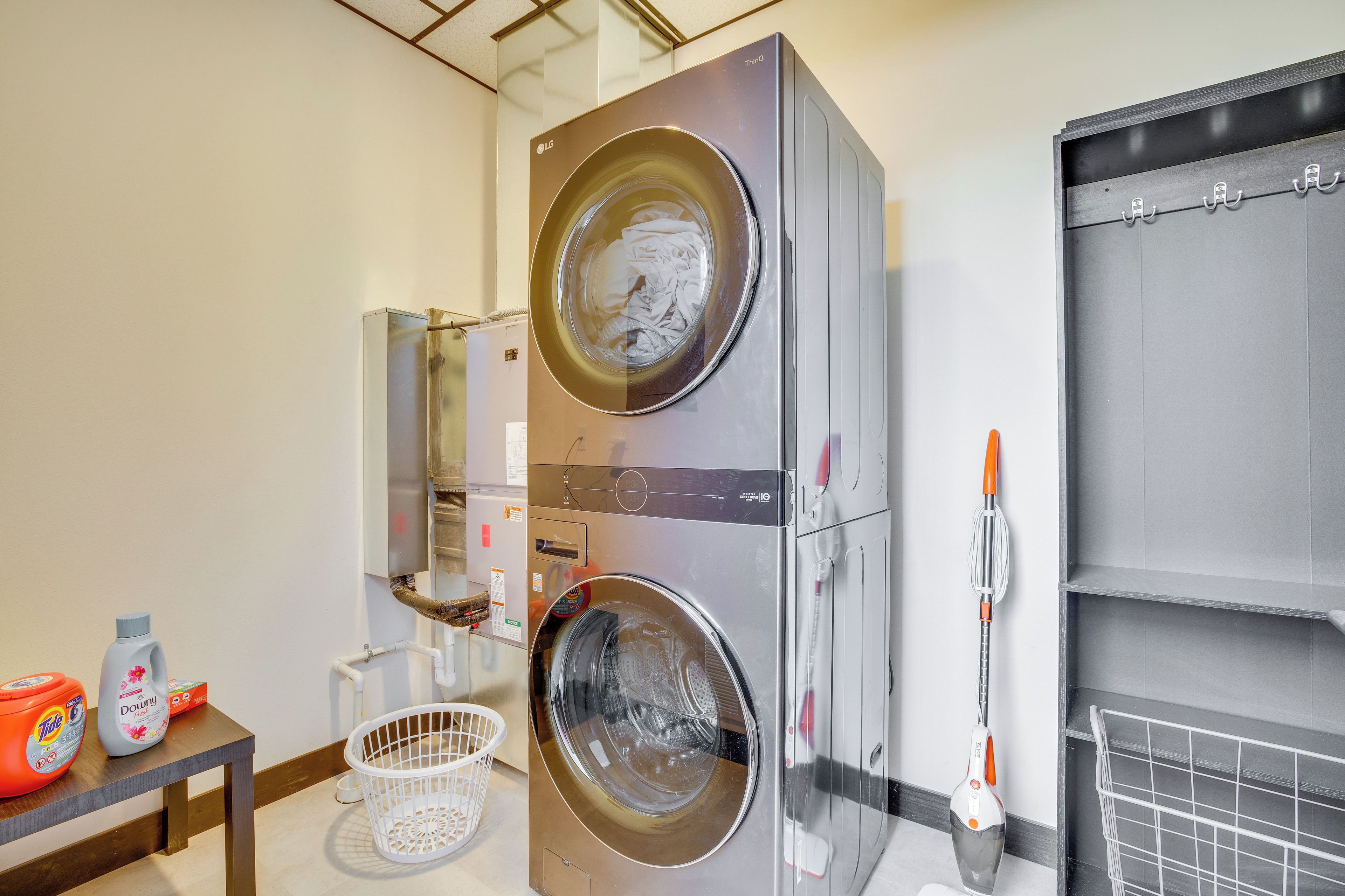 A modern laundry room with a double stainless steel washer and dryer, a white laundry basket, and a mop on a wall-mounted rack.