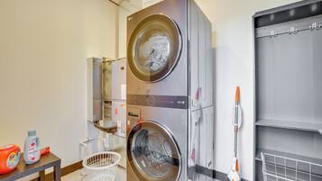 A modern laundry room with a double stainless steel washer and dryer, a white laundry basket, and a mop on a wall-mounted rack.