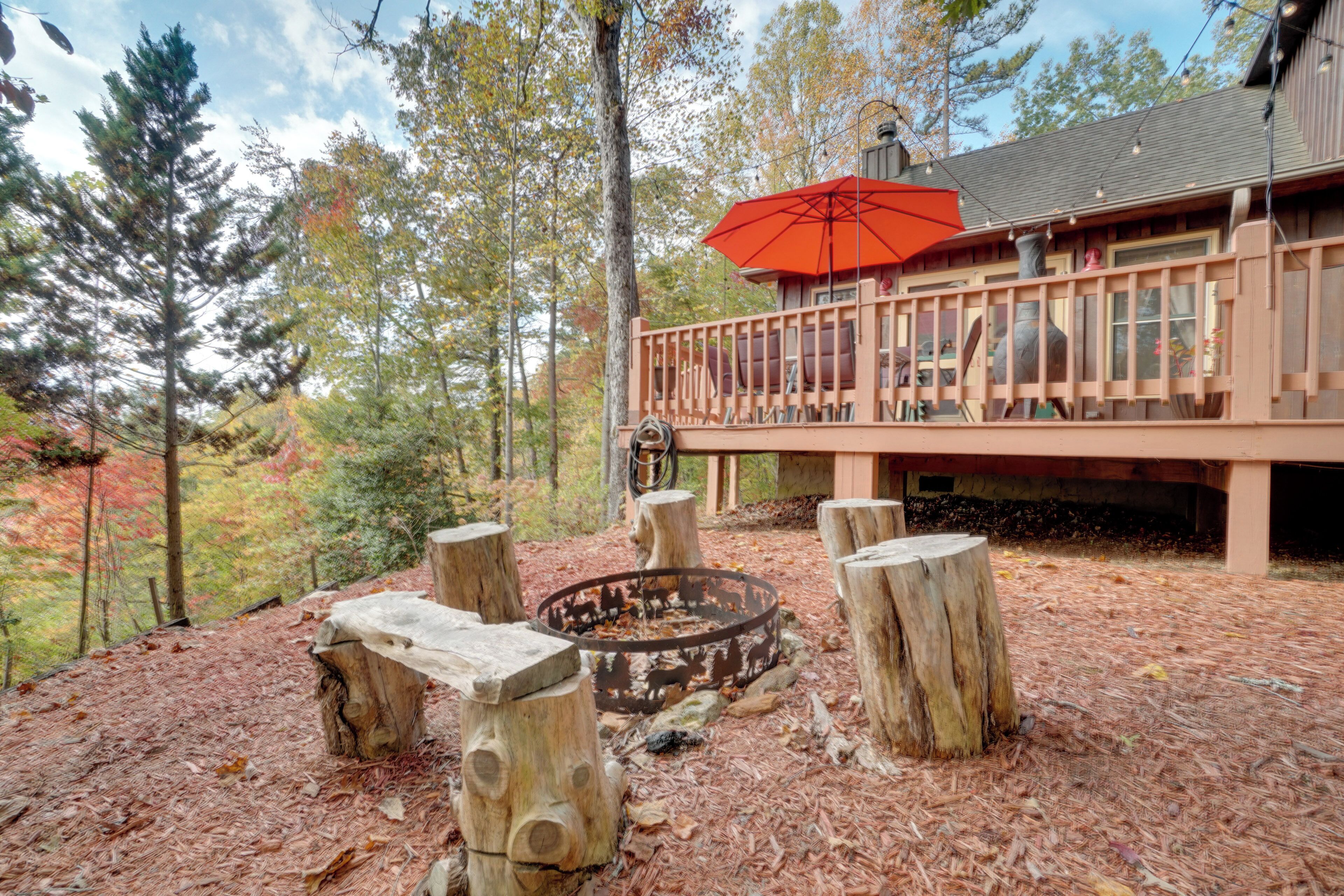 A wooden deck with a fire pit, surrounded by trees and a building with a red umbrella.