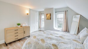 Ett sovrum med en säng, en byrå, en vägglampa, ett fönster med gardiner och en tavla på väggen.