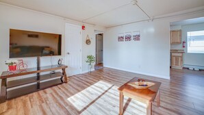 A spacious living room with hardwood flooring, a wooden coffee table, a flat-screen TV on a glass TV stand, and a door leading to another room. - Anchorage Apartment Rental w/ Mountain Views! (Anchorage)