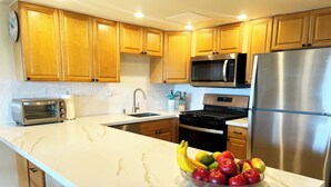 A kitchen with wooden cabinets, a stainless steel refrigerator, a microwave, a stove, a sink, and a bowl of fruit on the counter. - Bright Kihei Renovated Condo New AC Steps to Kamaole II Beach and Pool (Kihei)