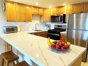 A kitchen with wooden cabinets, a stainless steel refrigerator, a microwave, a stove, a sink, and a bowl of fruit on the counter.