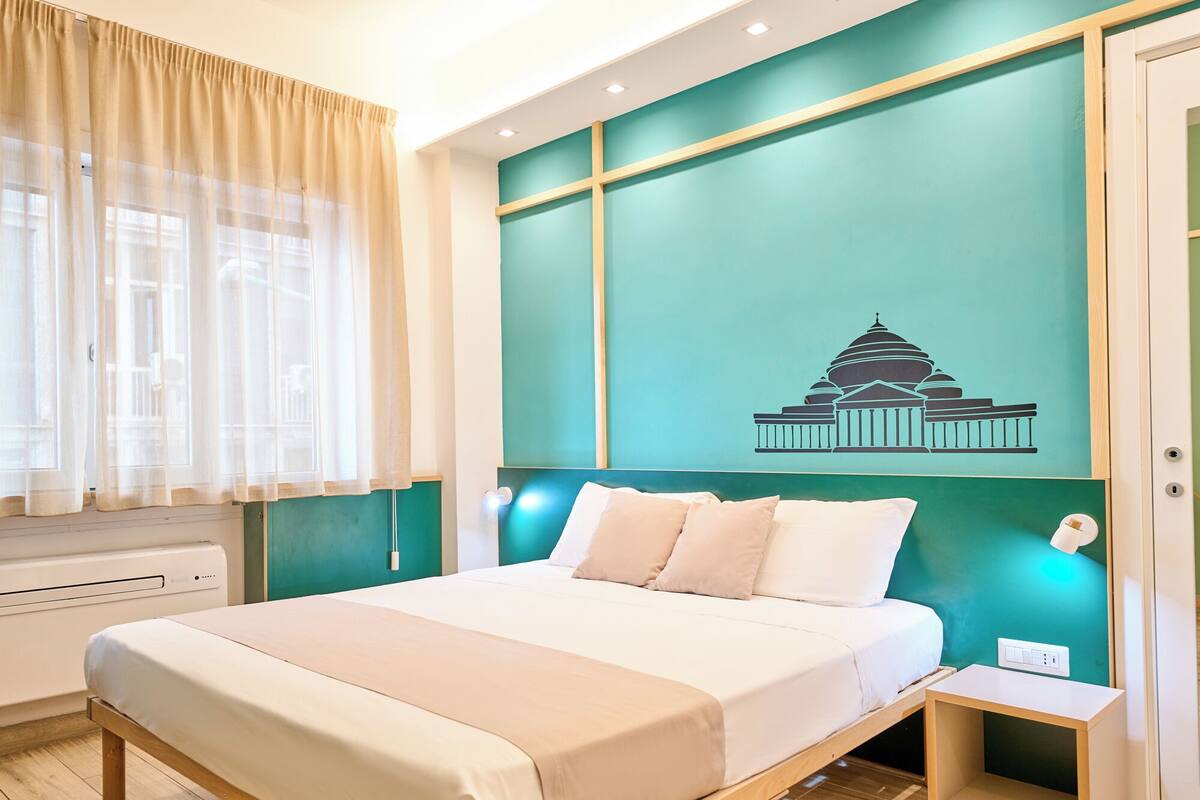 A hotel room with a bed, a nightstand, a wall-mounted lamp, and a window with sheer curtains.