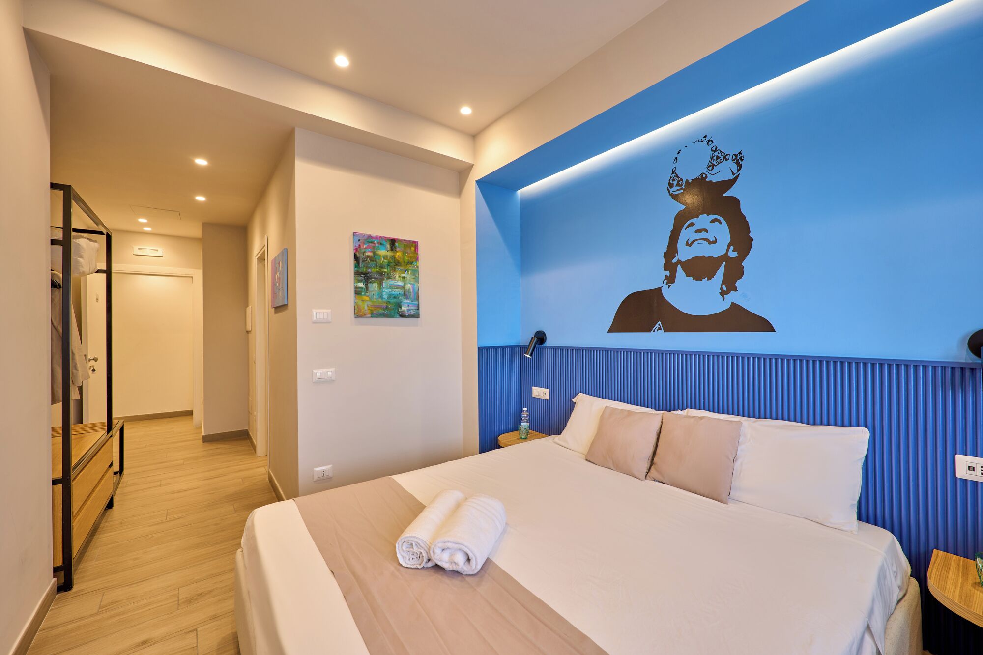 Una habitación de hotel con una cama, una pared con un mural, piso de madera y un pasillo.