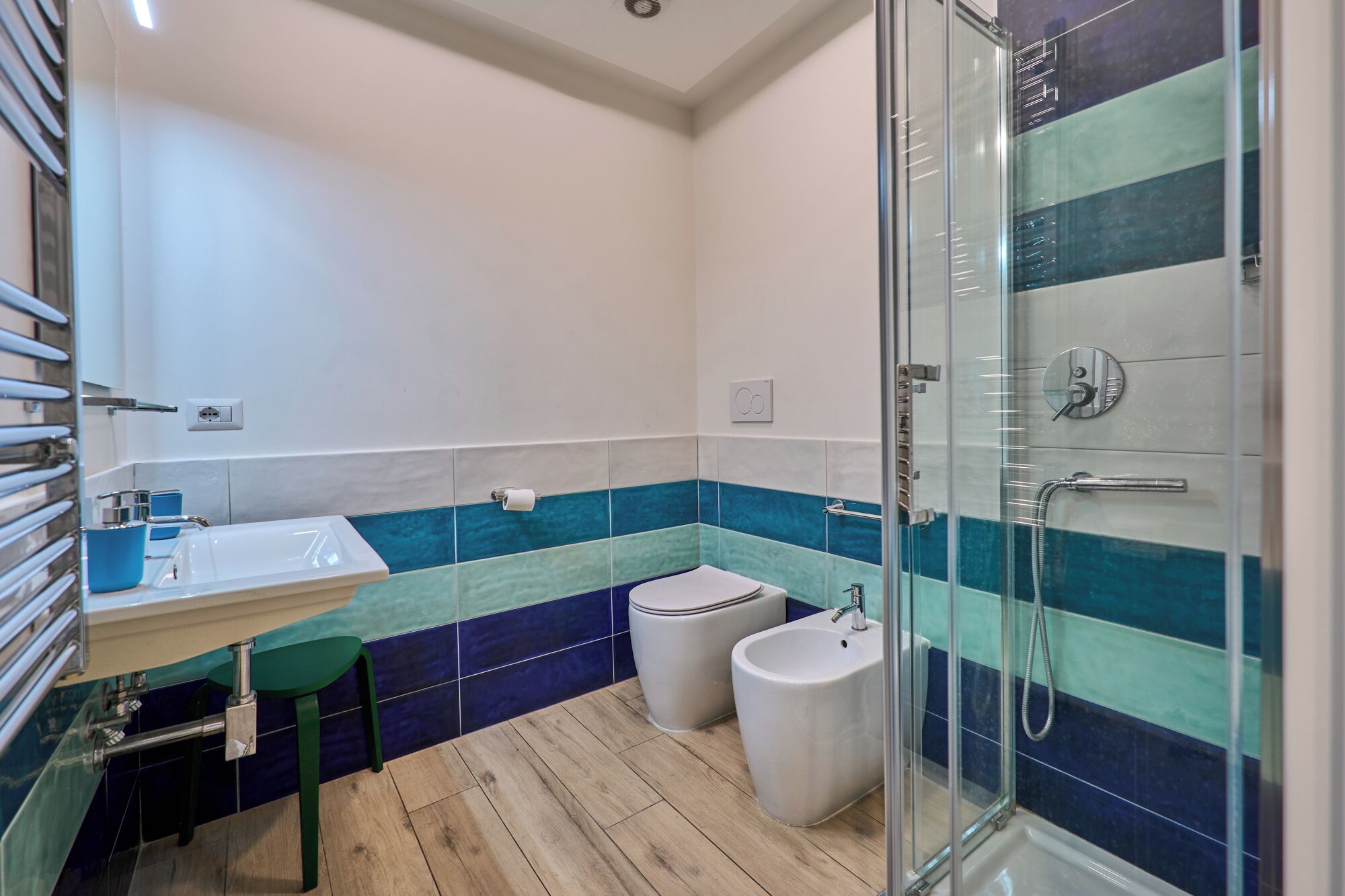Un baño moderno con cabina de ducha de vidrio, un lavabo con un taburete verde y dos inodoros.