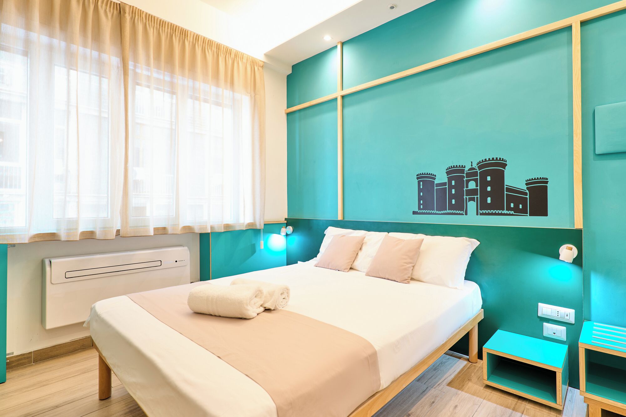 A hotel room with a bed, a window with sheer curtains, a wall-mounted air conditioner, and a teal-colored wall with a castle illustration.