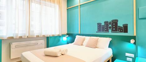 A hotel room with a bed, a window with sheer curtains, a wall-mounted air conditioner, and a teal-colored wall with a castle illustration.