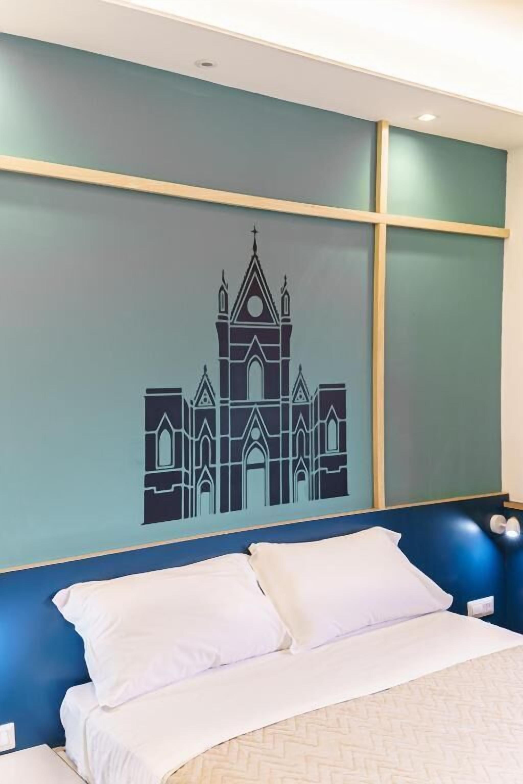 Habitación de hotel con una cama, almohadas blancas y una pared con un mural geométrico de una iglesia.