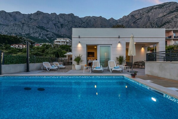 A swimming pool with lounge chairs, a modern building with a mountain view, and a patio area with outdoor furniture.