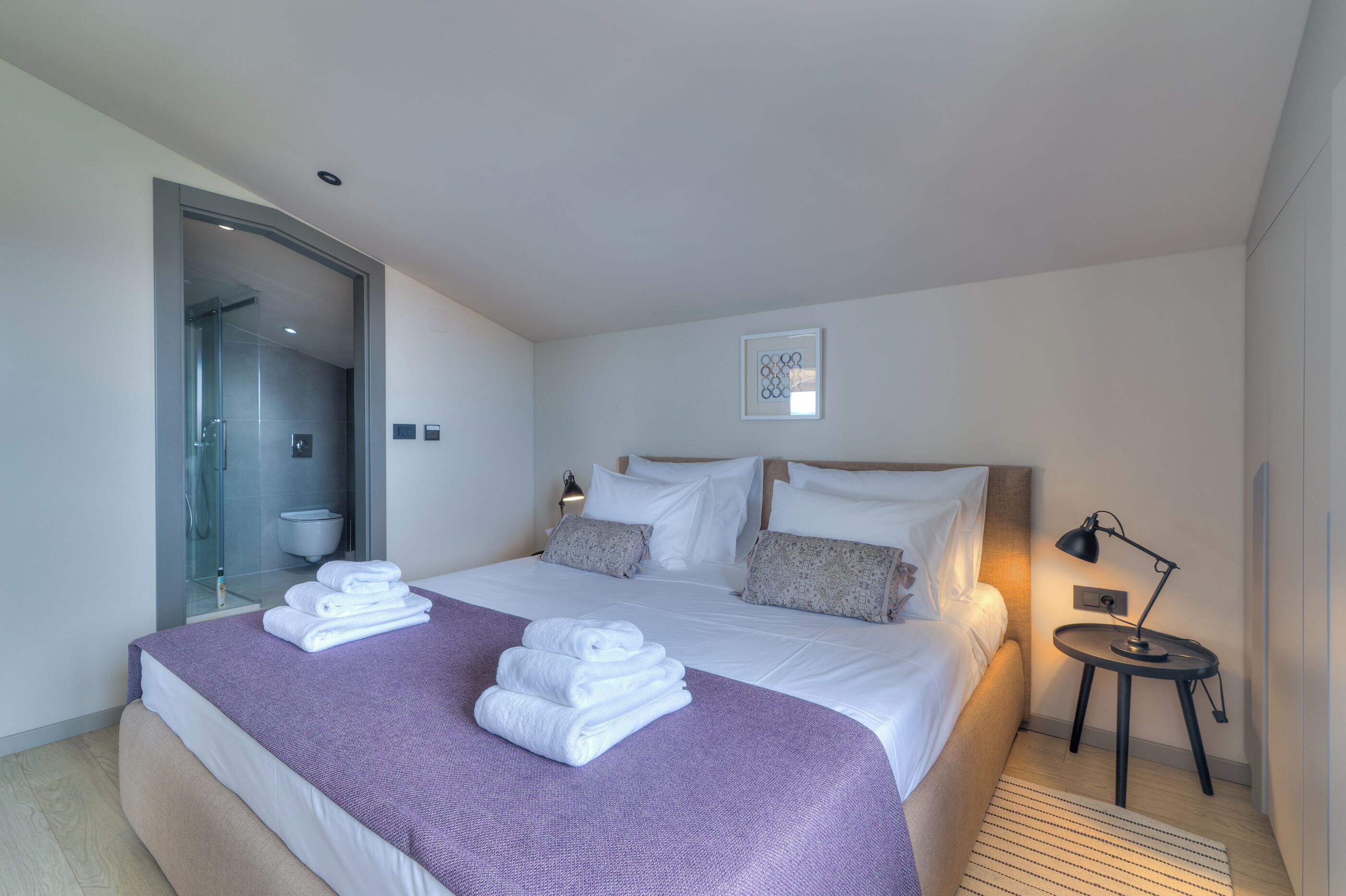 Une chambre d’hôtel moderne avec un grand lit, une douche en verre, une table de chevet avec une lampe et un tableau encadré au mur.