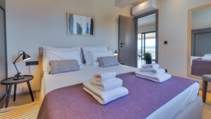 A modern hotel room with a large bed, a bedside table with a lamp, a painting on the wall, and a view through an open door.