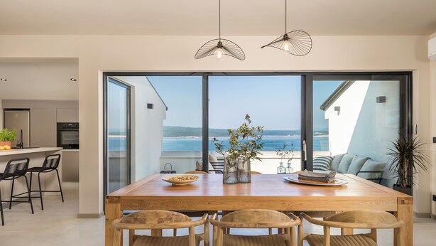 A modern dining area with a wooden table, four wicker chairs, and a view of the ocean through large glass doors.