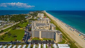Vue aérienne d’un hôtel situé sur la côte, composé de plusieurs bâtiments, d’un parking et d’une plage parsemée de parasols.