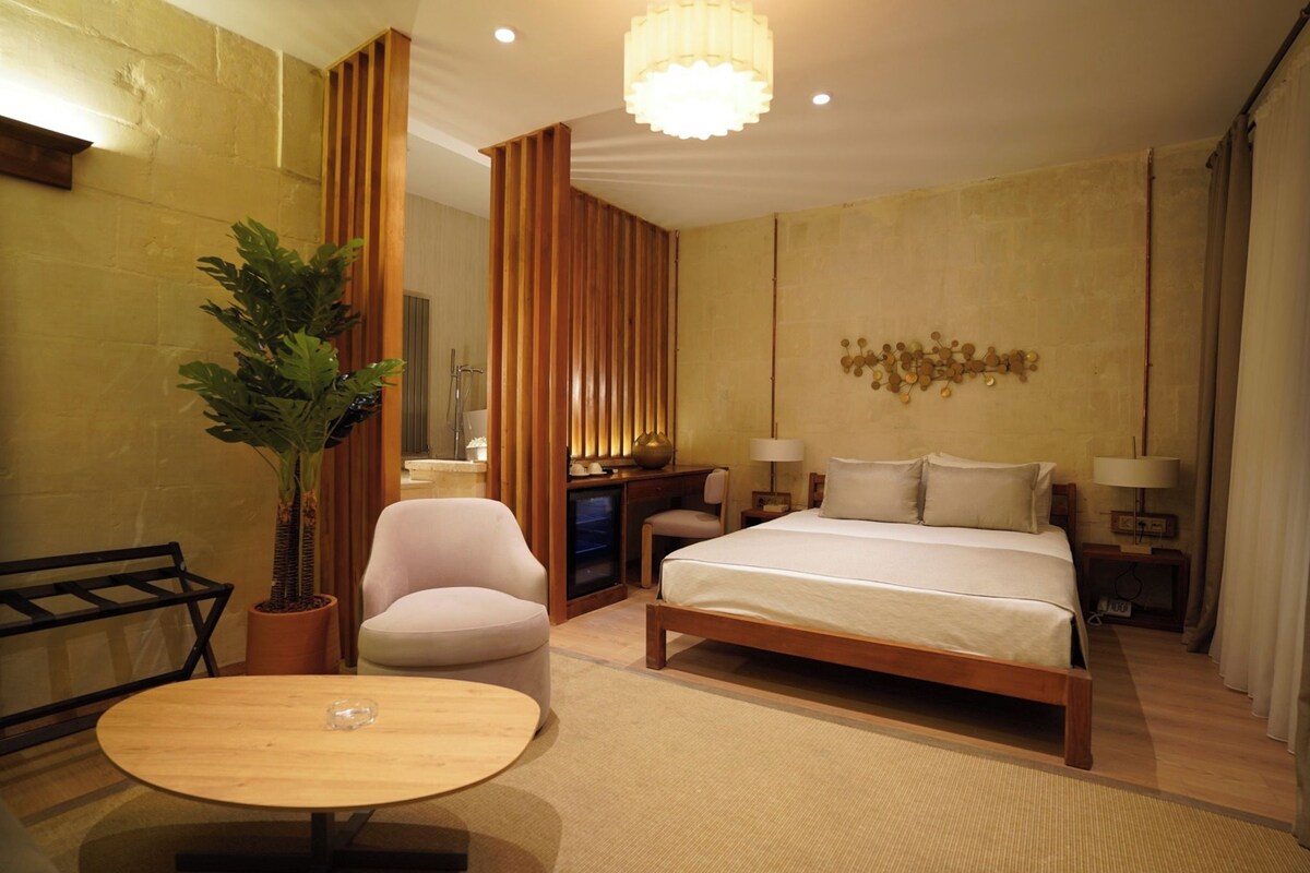A hotel room with a bed, a chair, a table, a potted plant, and a decorative wall piece.
